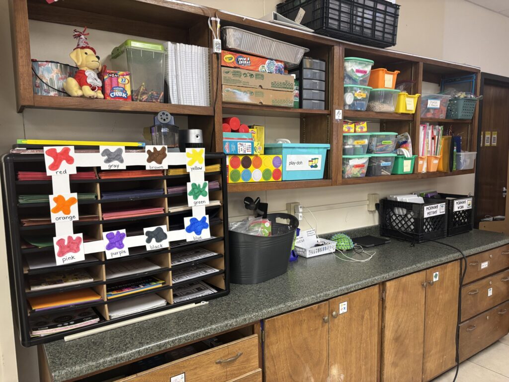 Classroom open shelving is organized in labeled plastic bins.