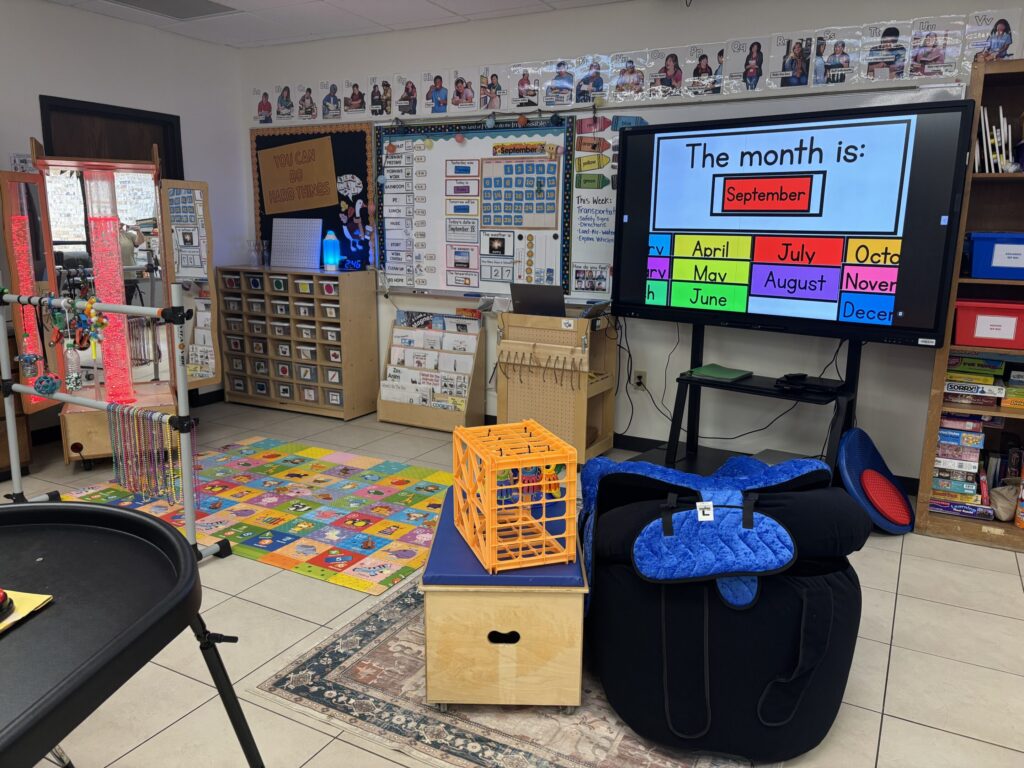 The front of the classroom is shown with a smart board. The calendar/morning meeting pieces are shown on the whiteboard behind. Adapted seating is in front of the board.