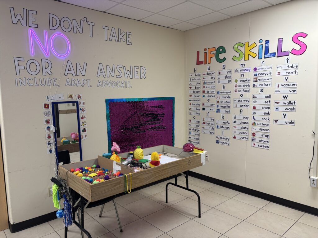 A sensory table sits in a corner with a life skills word wall, a sequin sensory wall, and a mirror.