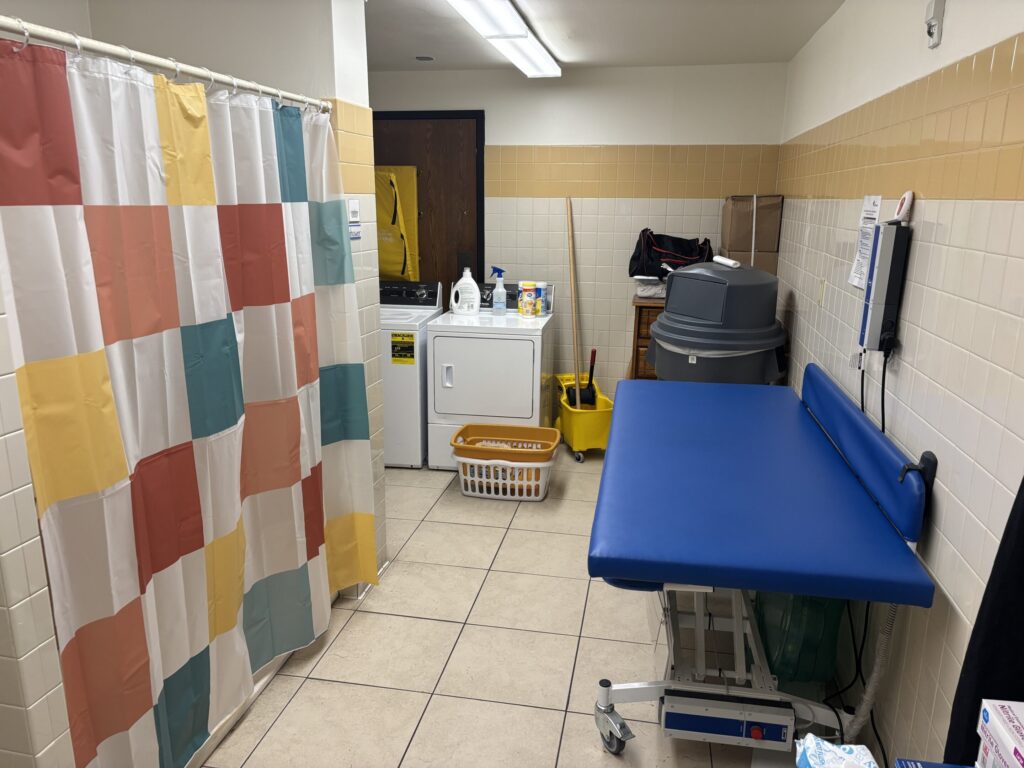 In a bathroom, a blue changing table is shown on the right side, and a colorful shower curtain hides a shower on the left. Washing machines and a trash can are shown in the background.