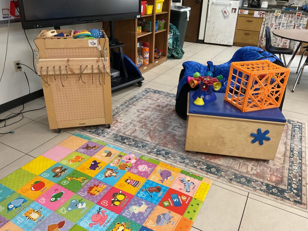 Furniture including an adaptive seating bean bag chair, a rolling stool, and a rolling cart are placed on a rug for student engagement in music. On the stool are. An adapted musical toy and a sensory crate with musical instruments hanging. On top of the cart are additional musical instruments.